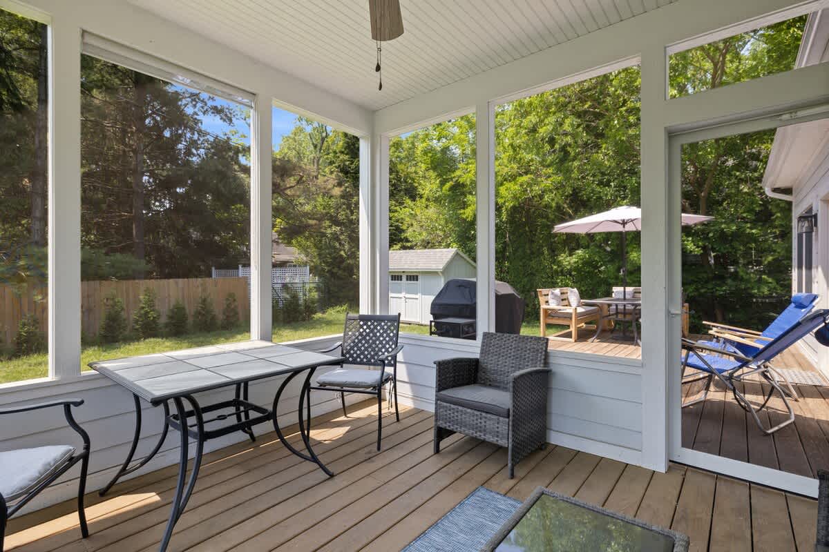 Another angle of the screened porch, showcasing a black metal dining set and a gray wicker armchair, with a view of the outdoor deck, grill, and a shed in the sunny backyard.
