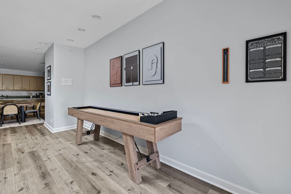 A light wood shuffleboard table with a black playing surface is positioned against a light gray wall adorned with three framed abstract artworks and a framed "Rules of Shuffleboard" poster.