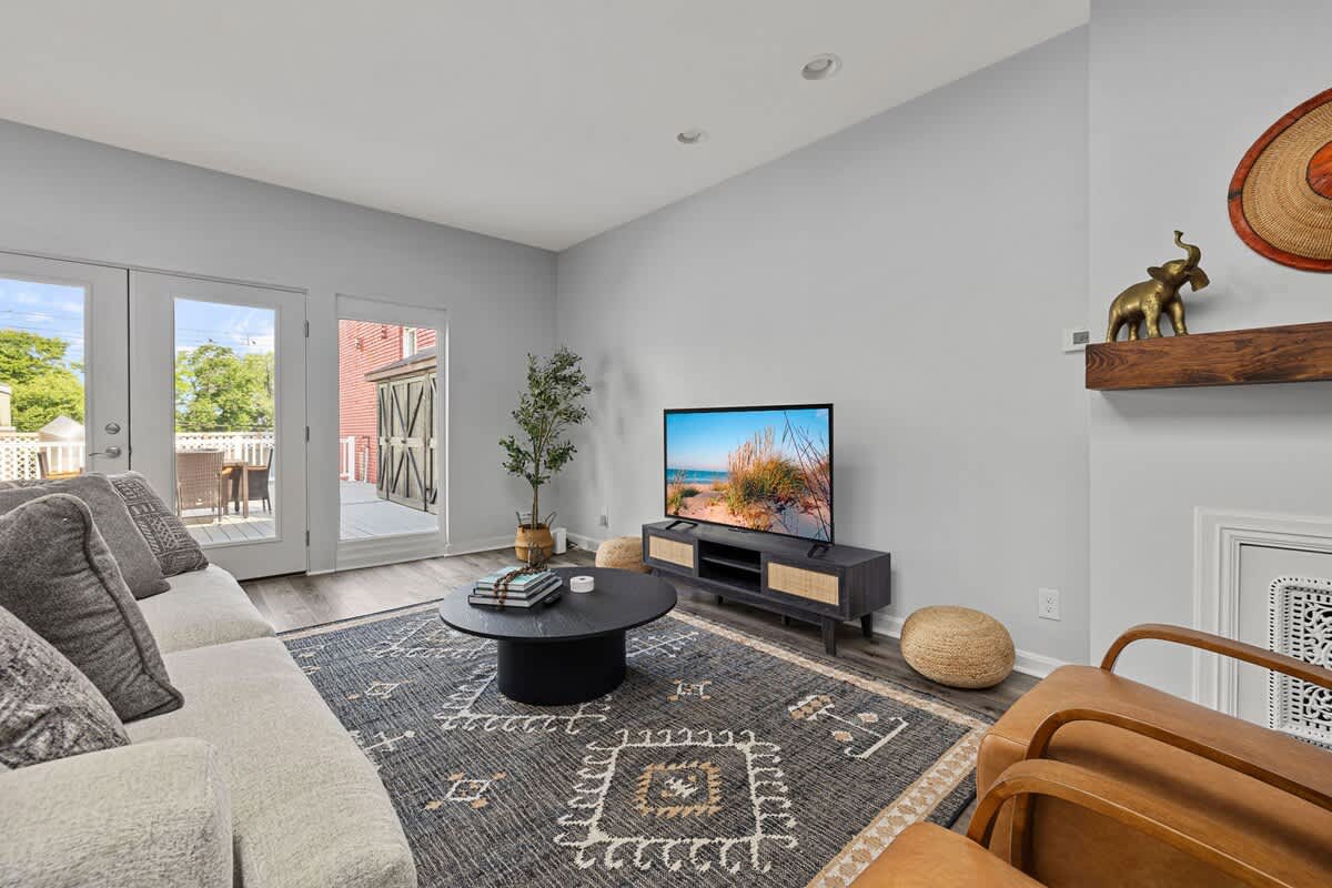 A cozy living room corner features a large flat-screen TV on a dark media console, two tan leather armchairs, and a black coffee table on a patterned rug, with decorative elements on a wall shelf.