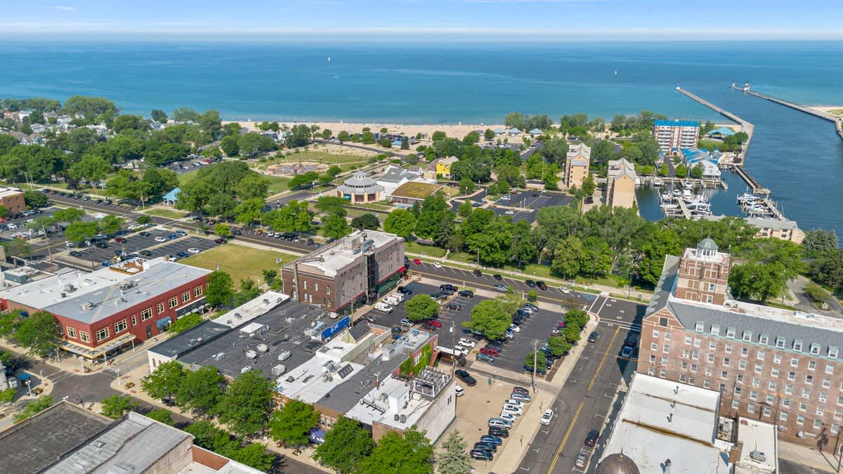 Another aerial perspective of the coastal town, emphasizing the close proximity of the urban structures to the sandy beach, marina, and the vast body of water. Green spaces and various parking areas are interspersed throughout the landscape.