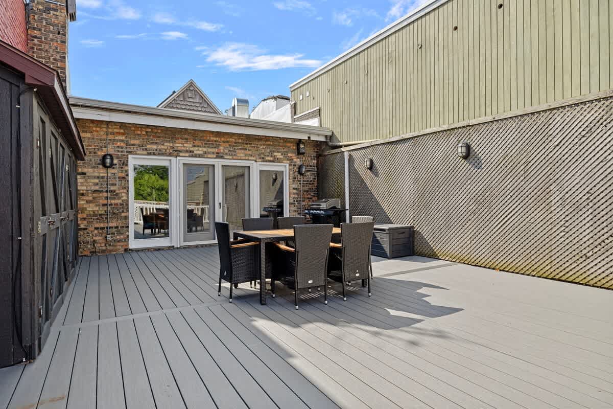 A spacious outdoor deck with gray composite decking, featuring a dark brown wicker dining set. The deck is adjacent to a brick building with large glass doors and a grill, and a light green building with slatted siding.