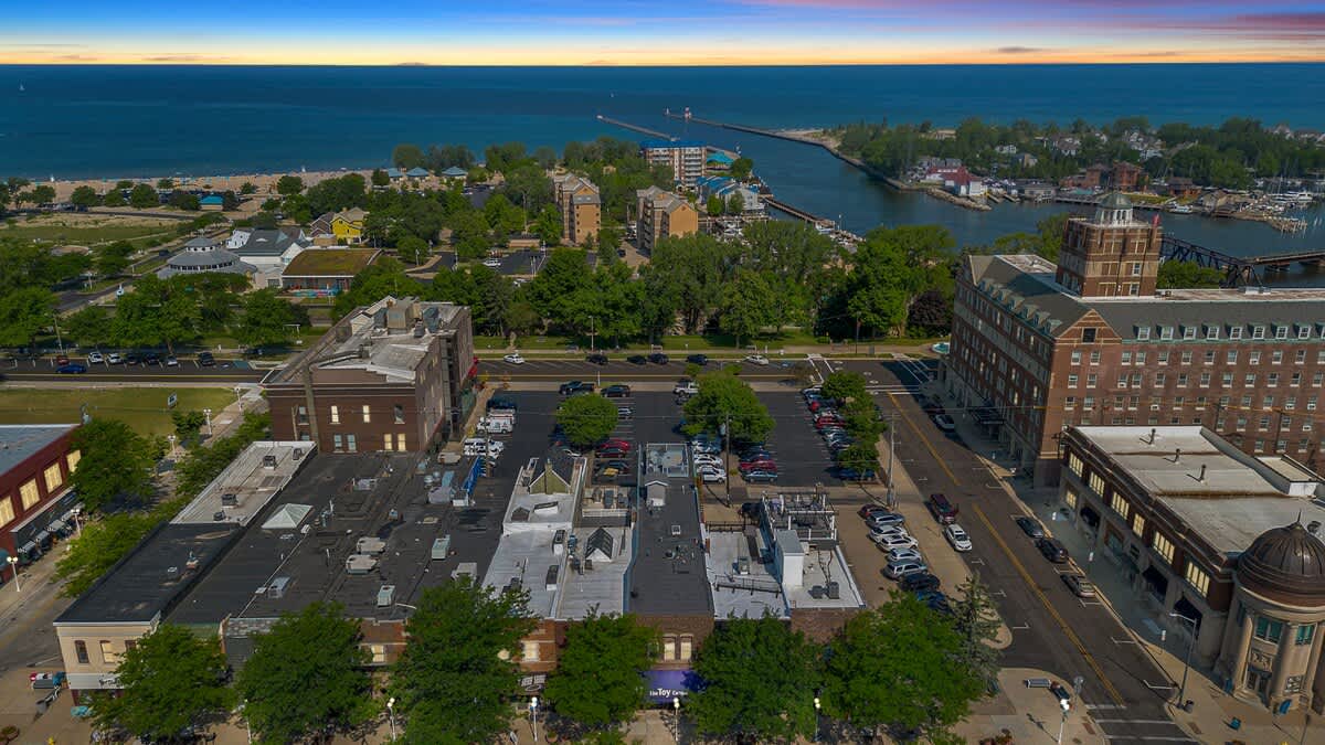 An aerial view captures a vibrant coastal town with a mix of commercial and residential buildings, parking lots, and streets, leading to a large body of water with a pier under a colorful sunset sky.