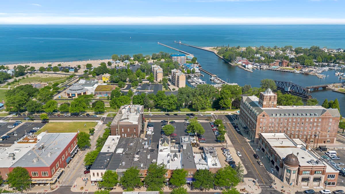 An expansive aerial view of a coastal town, featuring a mix of buildings, streets, and parking lots, with a large body of water (likely a lake or ocean) and a long pier extending into the distance. A sandy beach and a marina are also clearly visible.