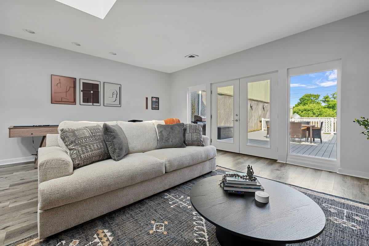 The bright living room features a light gray sofa with throw pillows, a black oval coffee table on a patterned rug, and French doors leading to an outdoor deck, with modern artwork on the wall.