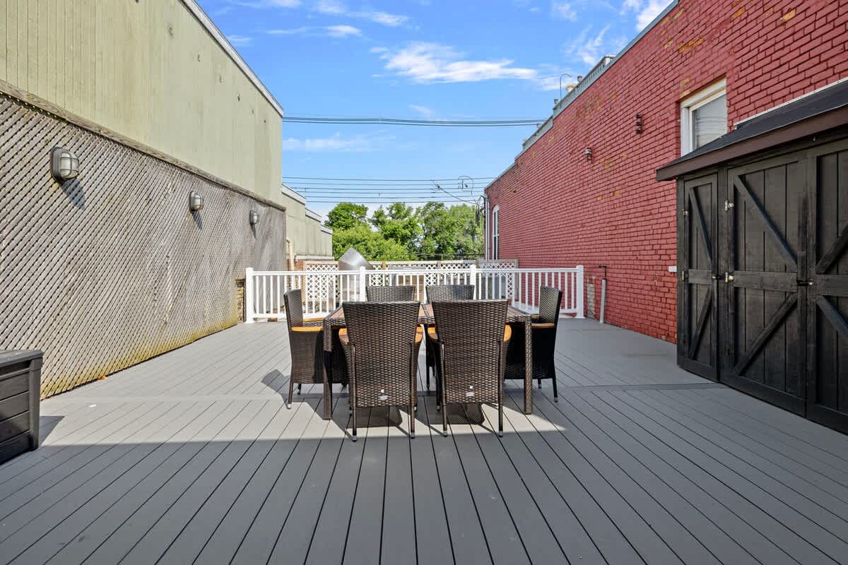 An outdoor deck with gray composite decking, featuring a dark brown wicker dining set with a rectangular table and eight chairs. The deck is enclosed by a white railing and flanked by a light green building and a red brick building with a black shed.