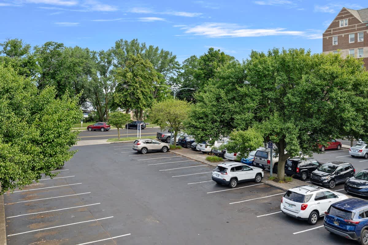 An elevated view of a large asphalt parking lot, neatly organized with numerous cars parked in marked spaces. The lot is surrounded by mature green trees, with various buildings visible in the background.
