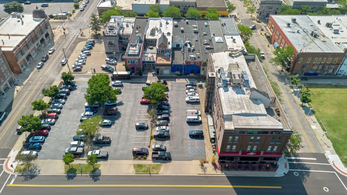 An aerial view looking down on a bustling street lined with multiple commercial buildings and a large, well-populated parking lot filled with cars. Mature trees provide shade along the parking lot, with other buildings visible in the background.