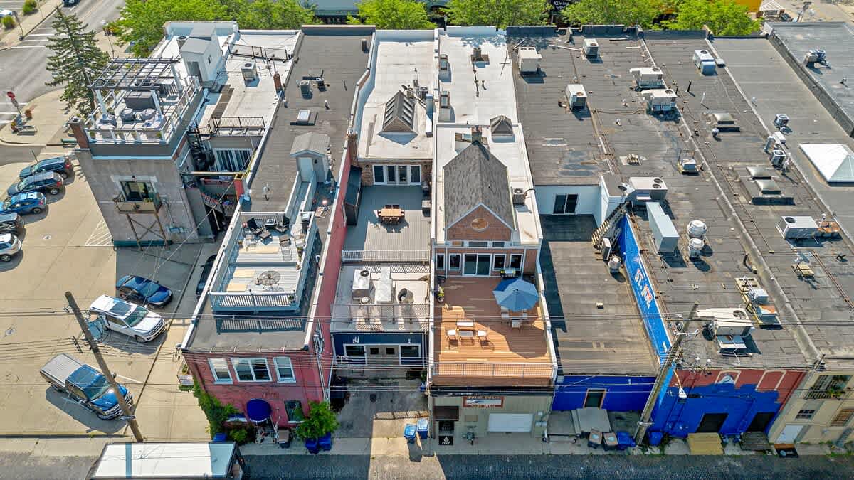A high aerial view showcasing the diverse rooftops of several buildings, some featuring inviting outdoor deck areas complete with patio furniture. A parking lot filled with cars is visible to the left, alongside various rooftop structures and skylights.