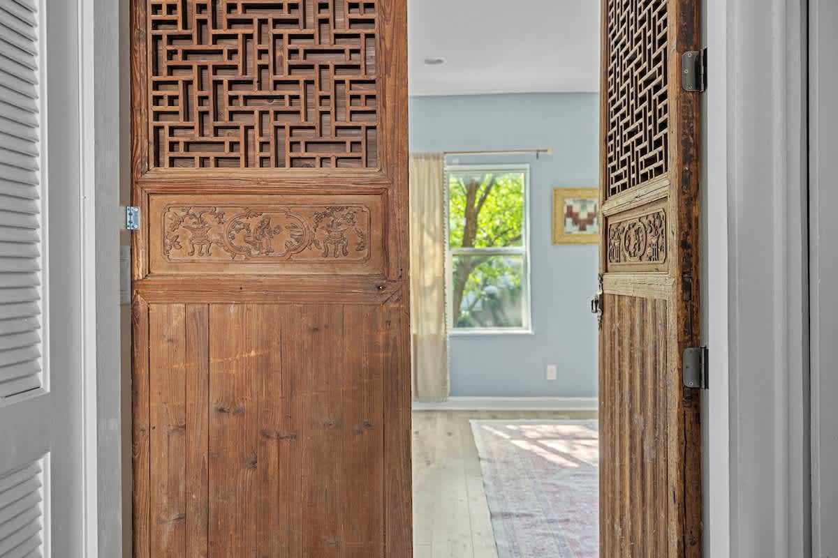 Ornate, dark wooden bi-fold doors with intricate carved patterns and geometric lattice work are open, revealing a glimpse of a light blue room with a window and a rug.