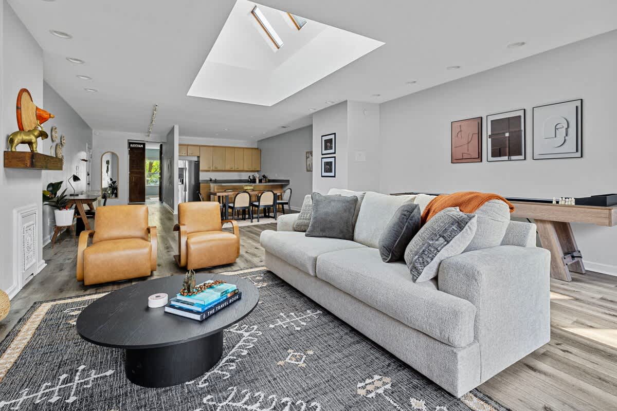 An expansive open-concept living area features a light gray sofa, two tan leather armchairs, and a black oval coffee table on a patterned rug, with a kitchen and dining area visible in the background.