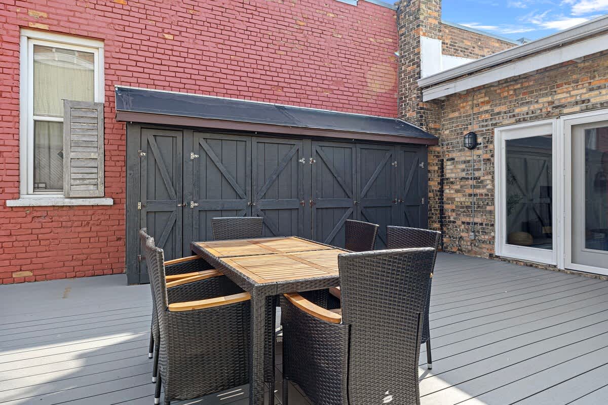 A close-up view of the outdoor deck, showcasing a dark brown wicker dining set with a wooden tabletop. In the background, there's a red brick building with a window and a large black shed with barn-style doors.