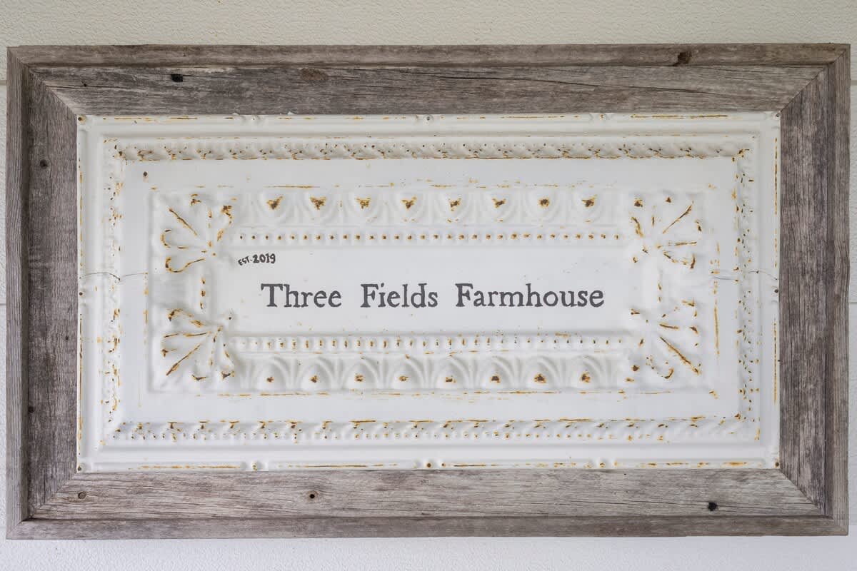 A rustic sign, 'Three Fields Farmhouse est. 2019,' is displayed in a distressed wooden frame, featuring a white embossed metal background with gold accents.