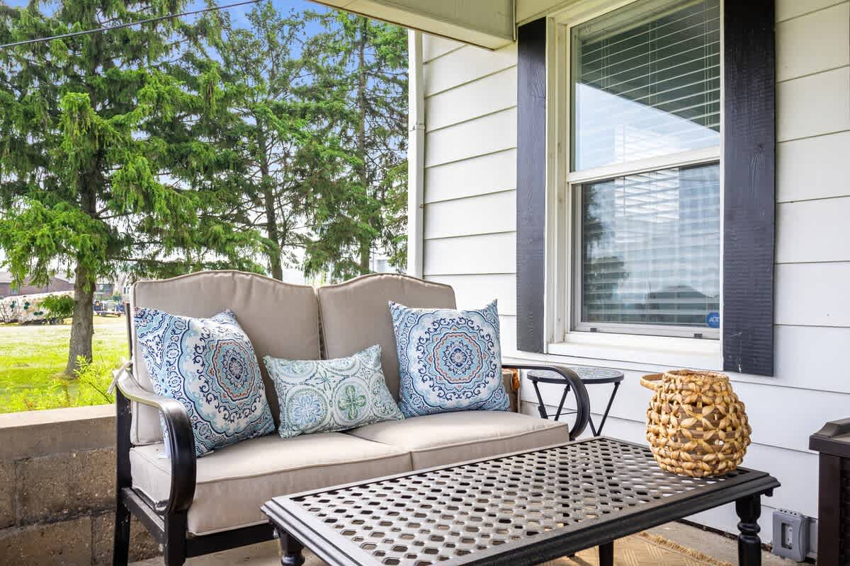 A cozy section of the covered porch features an outdoor sofa with decorative pillows and a metal coffee table, framed by a window with dark shutters.