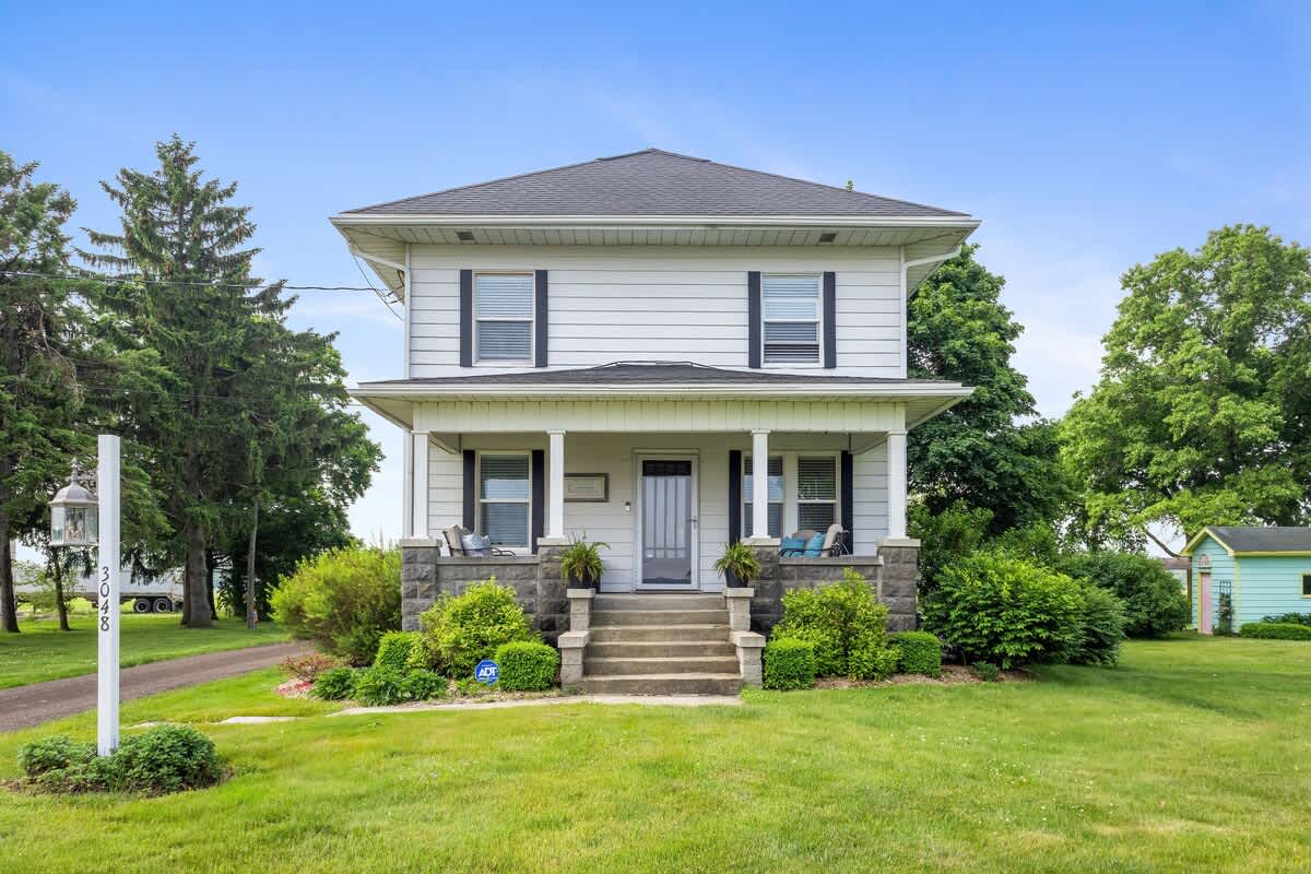 The front facade of a two-story white farmhouse with dark shutters, a covered porch supported by stone pillars, and a well-manicured green lawn.