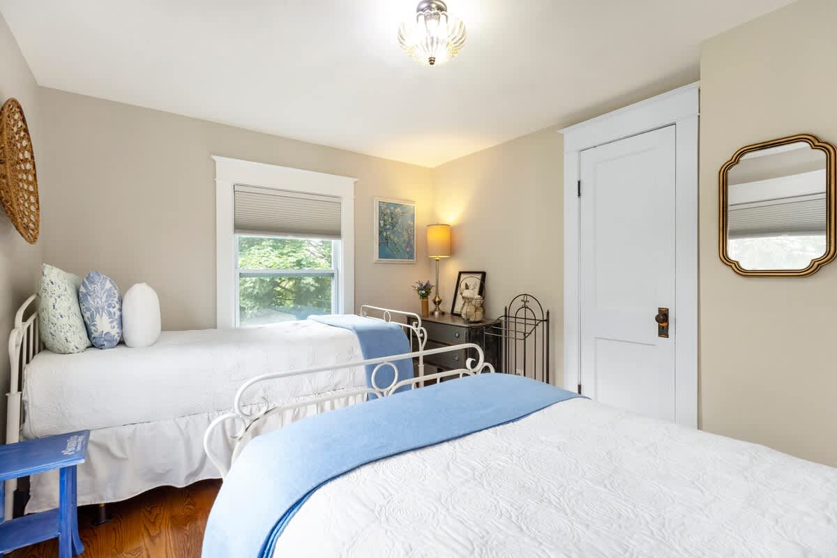 View of a bedroom with two white metal-frame twin beds, light beige walls, and hardwood floors. A small nightstand with a lamp sits between the beds, and a decorative mirror hangs near a white door.