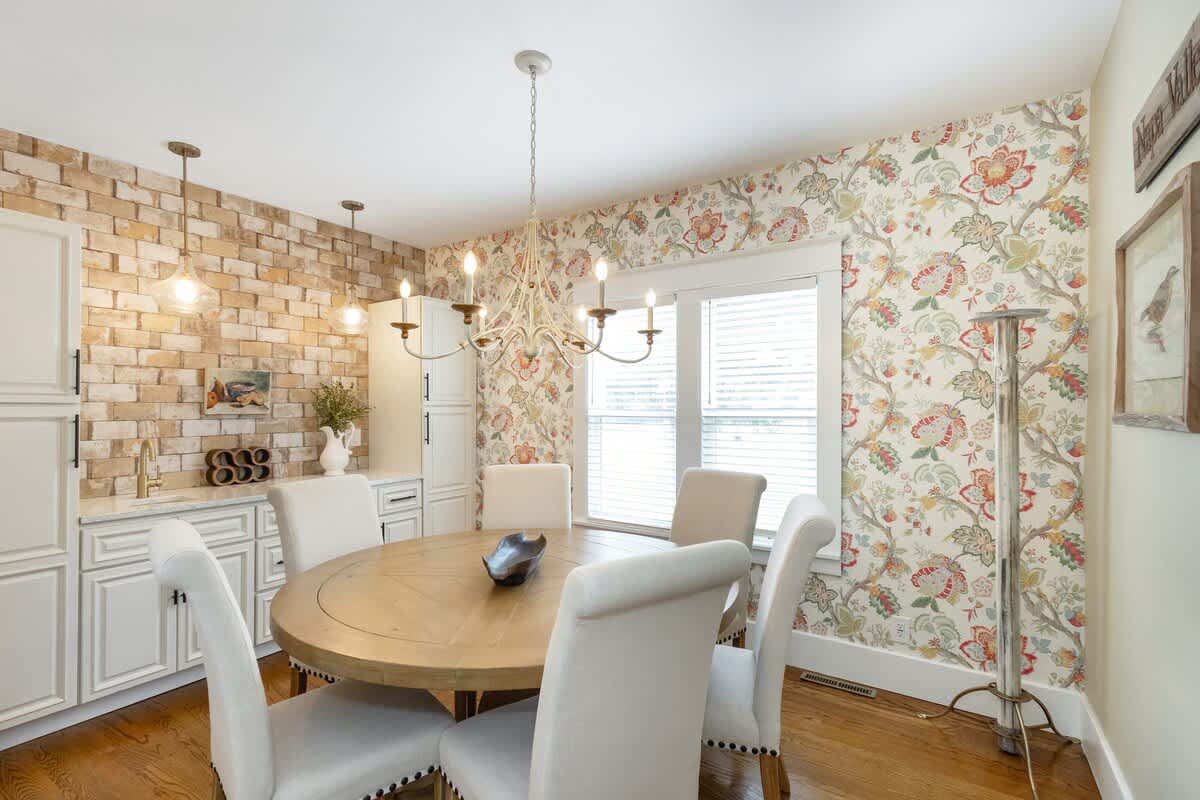 Elegant dining room with a round wooden table, four upholstered chairs, and a vibrant floral wallpaper accent wall. A built-in wet bar with brick backsplash and white cabinetry is visible on the left.