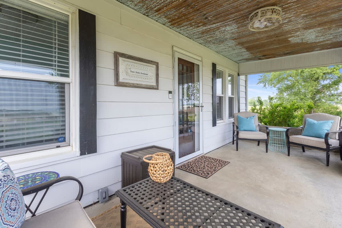 A cozy porch area features two armchairs with blue pillows, a small side table, and a black metal coffee table. The white siding of the house, a front door, and a decorative sign are visible.