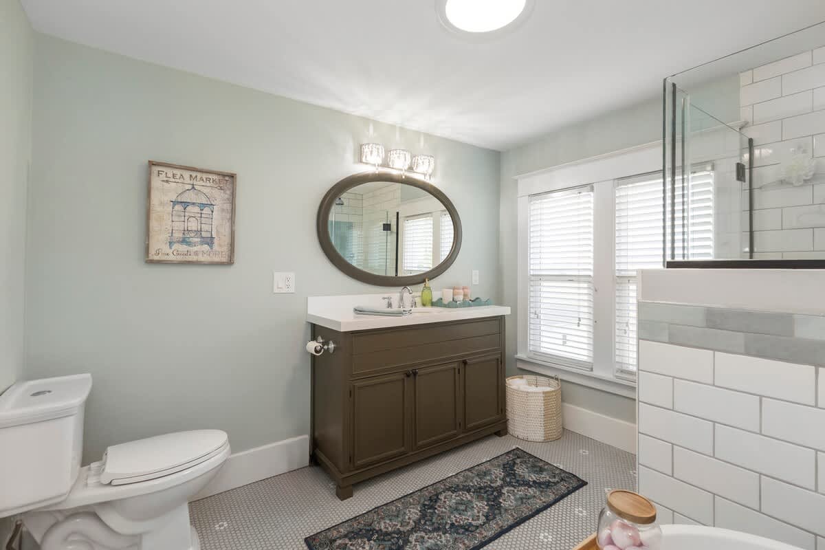Bright bathroom with light blue walls, a dark green vanity with an oval mirror, a toilet, and a glass-enclosed shower. Gray hexagonal floor tiles and a patterned rug complete the look.
