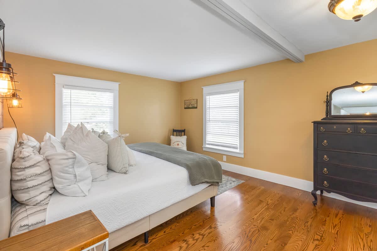 Bright bedroom with a large bed, light yellow walls, and polished hardwood floors. A dark dresser with a mirror stands against the far wall, and two windows with blinds let in light.