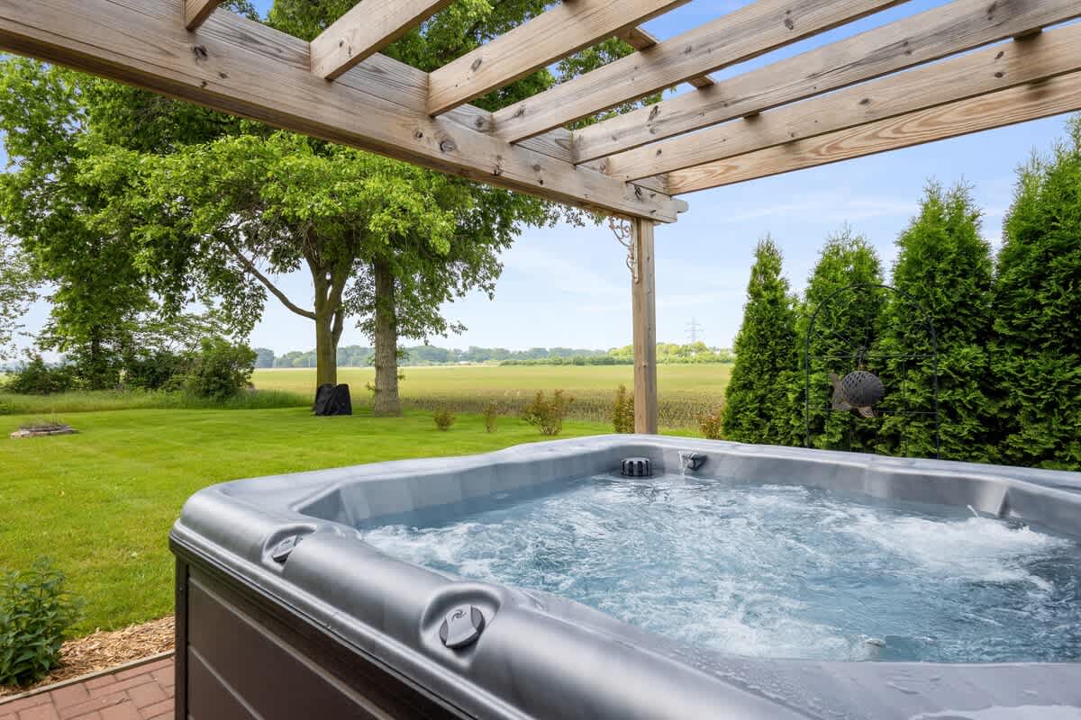 Outdoor hot tub with bubbling water, situated on a patio under a wooden pergola. Lush green grass, trees, and a distant field create a serene natural backdrop.