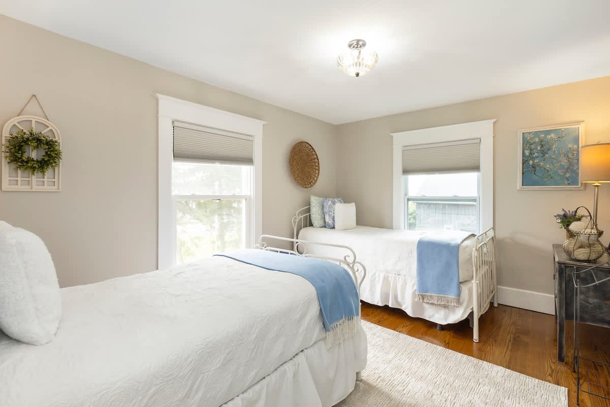 Cozy bedroom with two white metal-frame twin beds, each with white bedding and a light blue throw. Light beige walls, hardwood floors, and a light-colored rug create a calm space.