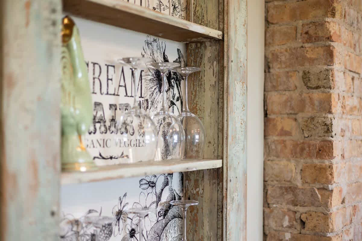 Close-up of a distressed, light green wooden shelving unit with decorative paper backing, displaying wine glasses and a ceramic bird figurine, positioned next to an exposed brick wall.