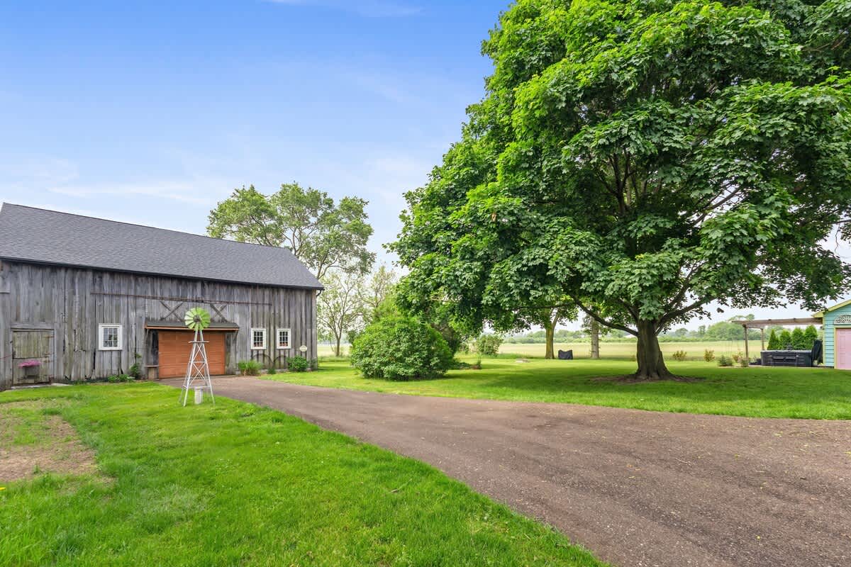 A long gravel driveway leads past a rustic wooden barn with a dark roof and a decorative windmill. A large, mature green tree dominates the right side of the frame, with expansive green lawn and fields beyond.