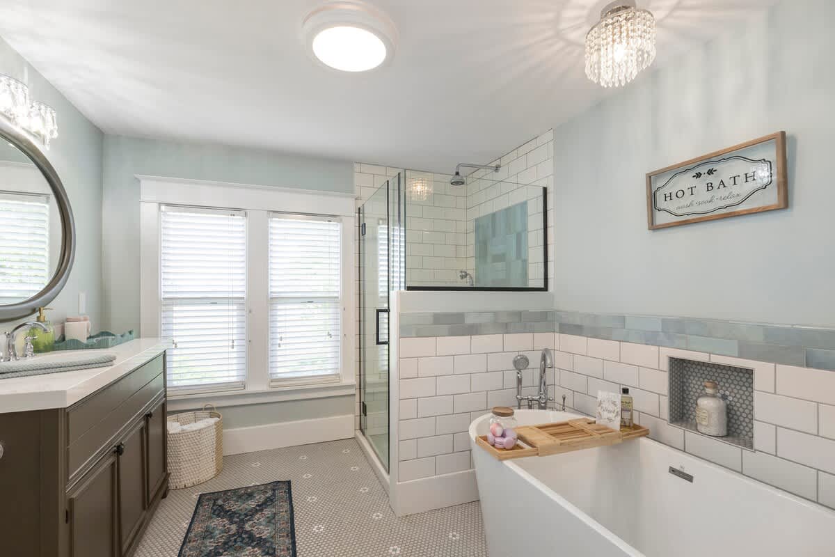 Spacious bathroom featuring a white freestanding bathtub, a glass-enclosed shower with subway tiles, and a dark green vanity. Light blue walls and gray hexagonal floor tiles create a serene atmosphere.