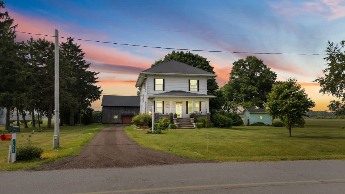 The charming white farmhouse with black shutters and a dark roof stands against a vibrant sunset sky. A gravel driveway leads to the front porch, flanked by mature trees and a well-maintained green lawn.