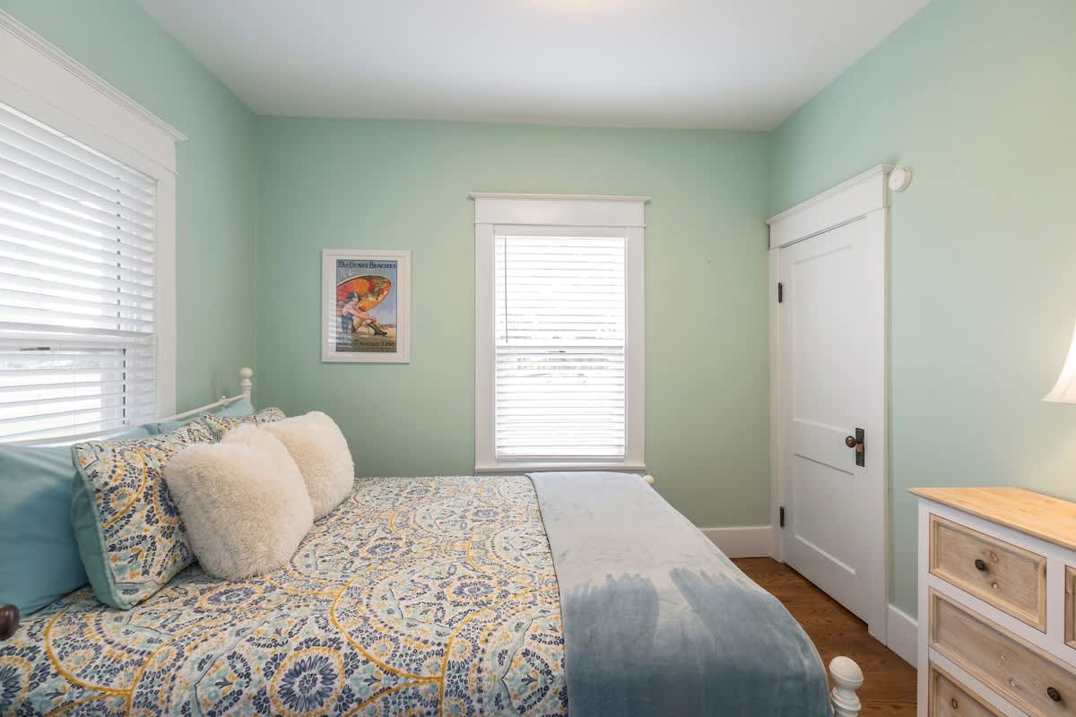 Inviting bedroom with light green walls, a queen-sized bed featuring a blue and yellow patterned duvet and fluffy white pillows. A large window with white blinds brightens the room.