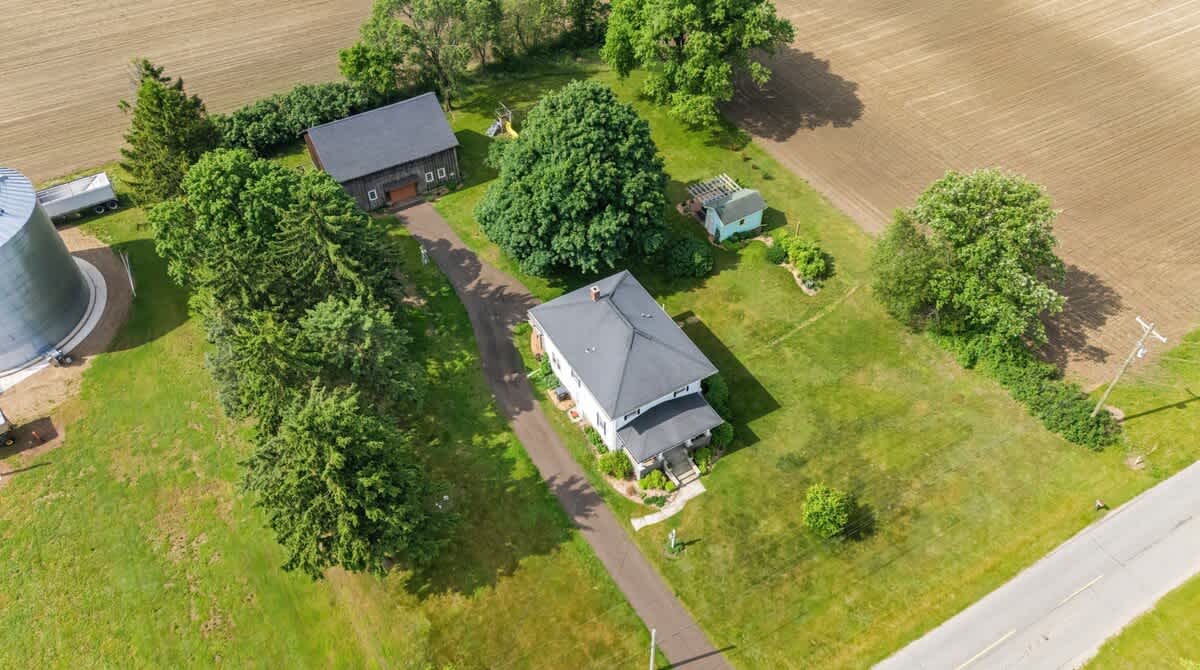 An aerial view showcases the white farmhouse with a dark roof, a long driveway, and a large barn surrounded by lush green lawns and mature trees. Agricultural fields border the property, highlighting its rural setting.