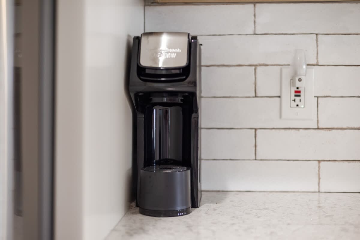 A black single-serve coffee maker sits on a white quartz countertop against a white subway tile backsplash, next to a white electrical outlet with a nightlight.