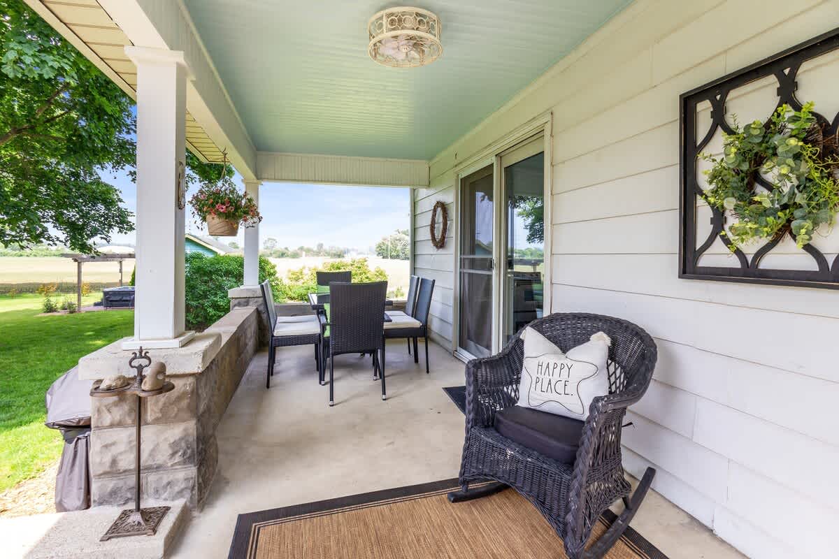 A spacious covered porch features an outdoor dining set and a wicker rocking chair with a 'Happy Place' pillow, offering a view of the expansive yard.