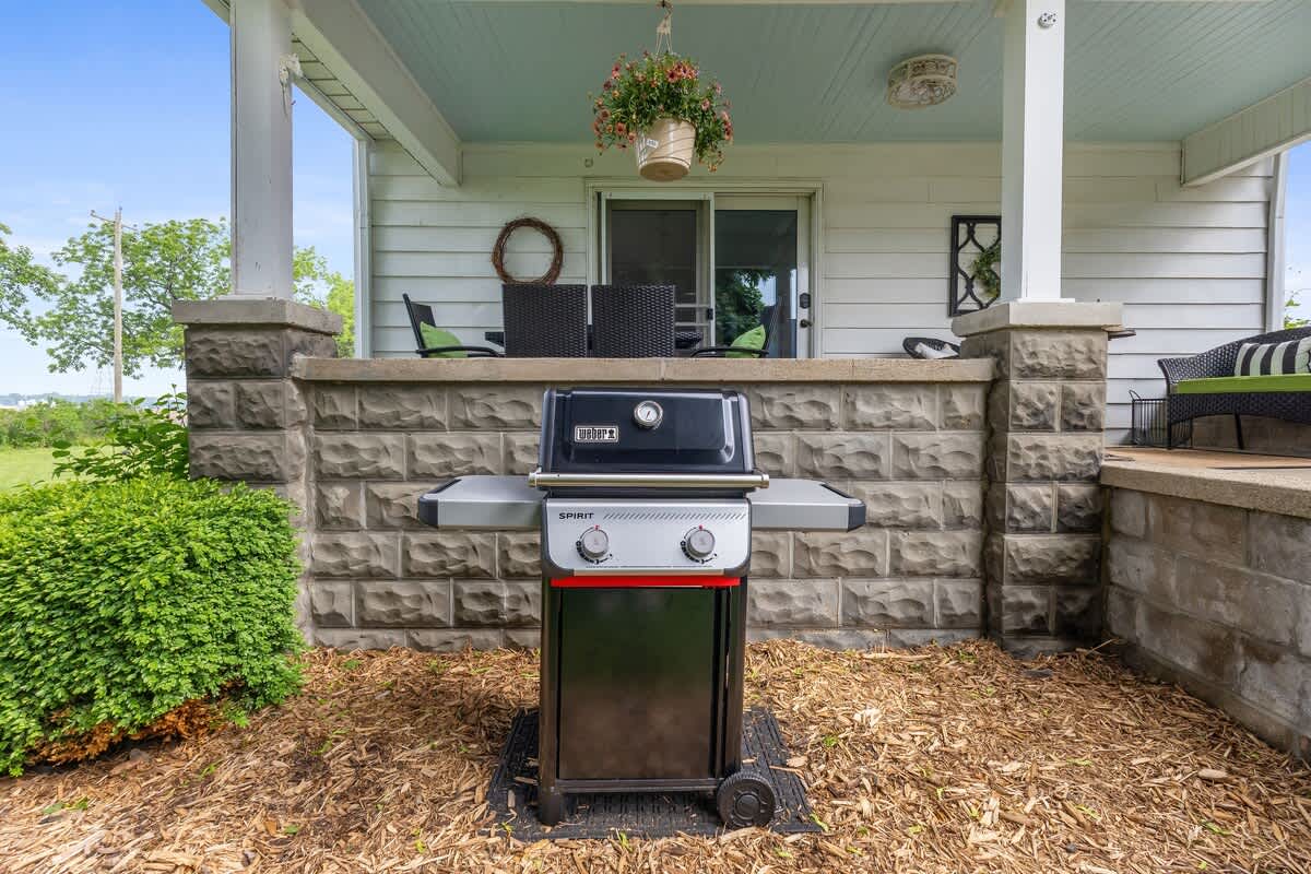 A black and silver Weber grill sits on a mat on a wood chip ground, in front of a stone-faced porch wall. The porch has white columns, a light blue ceiling, and outdoor seating visible in the background.