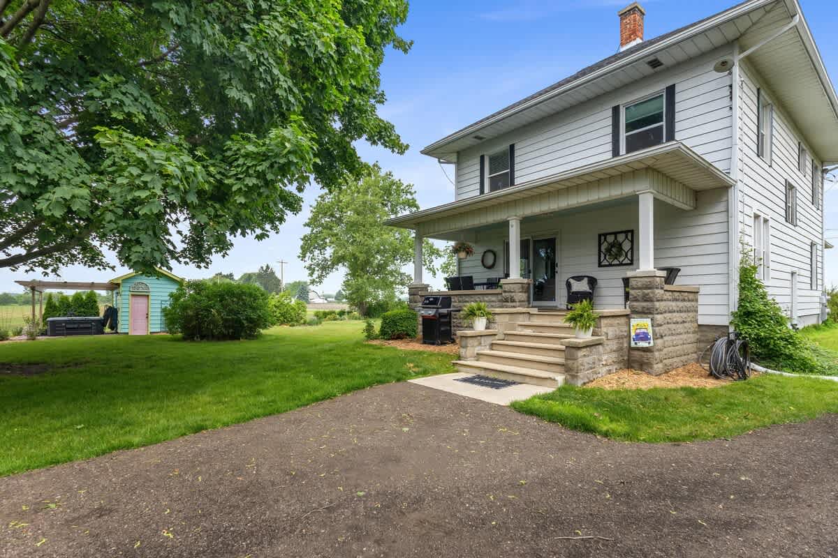 The side view of the white farmhouse shows a gravel driveway leading to a covered porch with a grill, overlooking a large grassy yard and the colorful shed in the distance.
