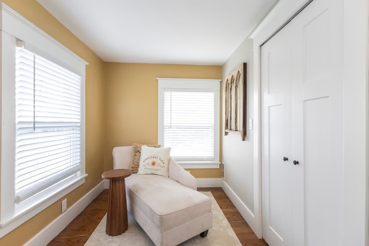 Cozy sitting area, part of bedroom-1, with light yellow walls, hardwood floors, and a cream-colored chaise lounge next to a small wooden side table. Two windows with white blinds provide natural light.