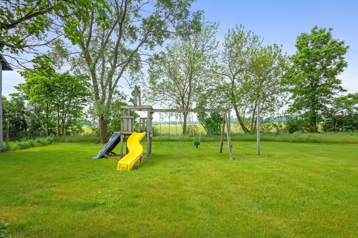 A children's playset with yellow and black slides and swings is set in a spacious, well-maintained green lawn, surrounded by trees and open fields in the distance.