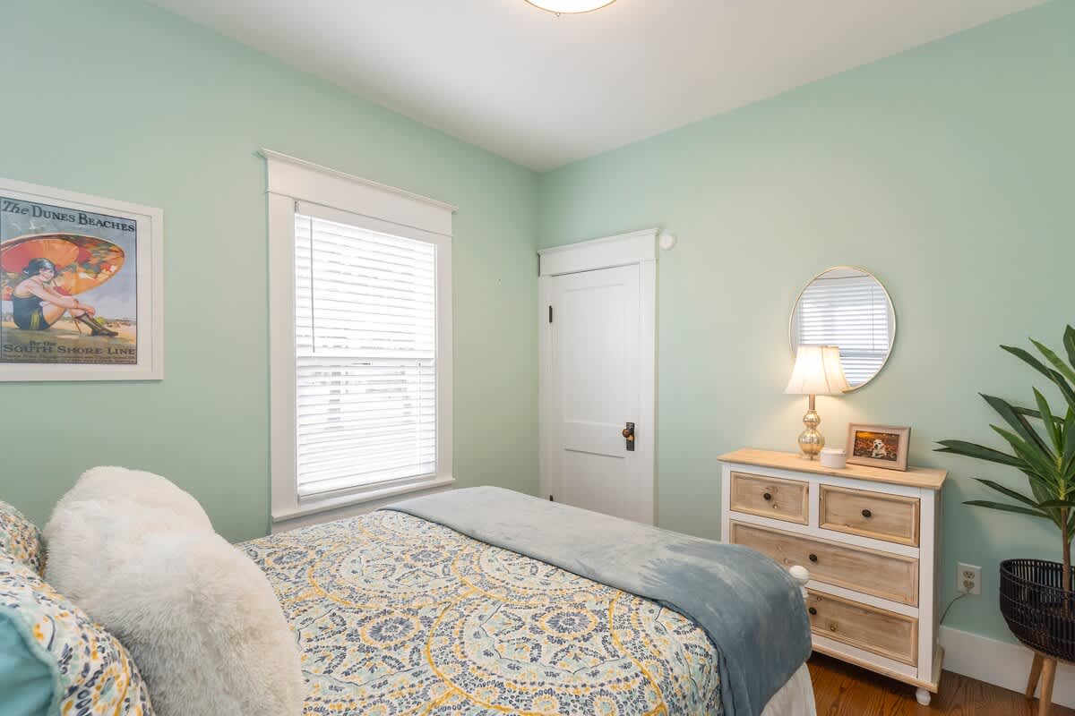 Bright bedroom with light green walls and a queen-sized bed with a colorful duvet. A rustic wooden dresser with a lamp and a round mirror sits beside the bed, near a large window.
