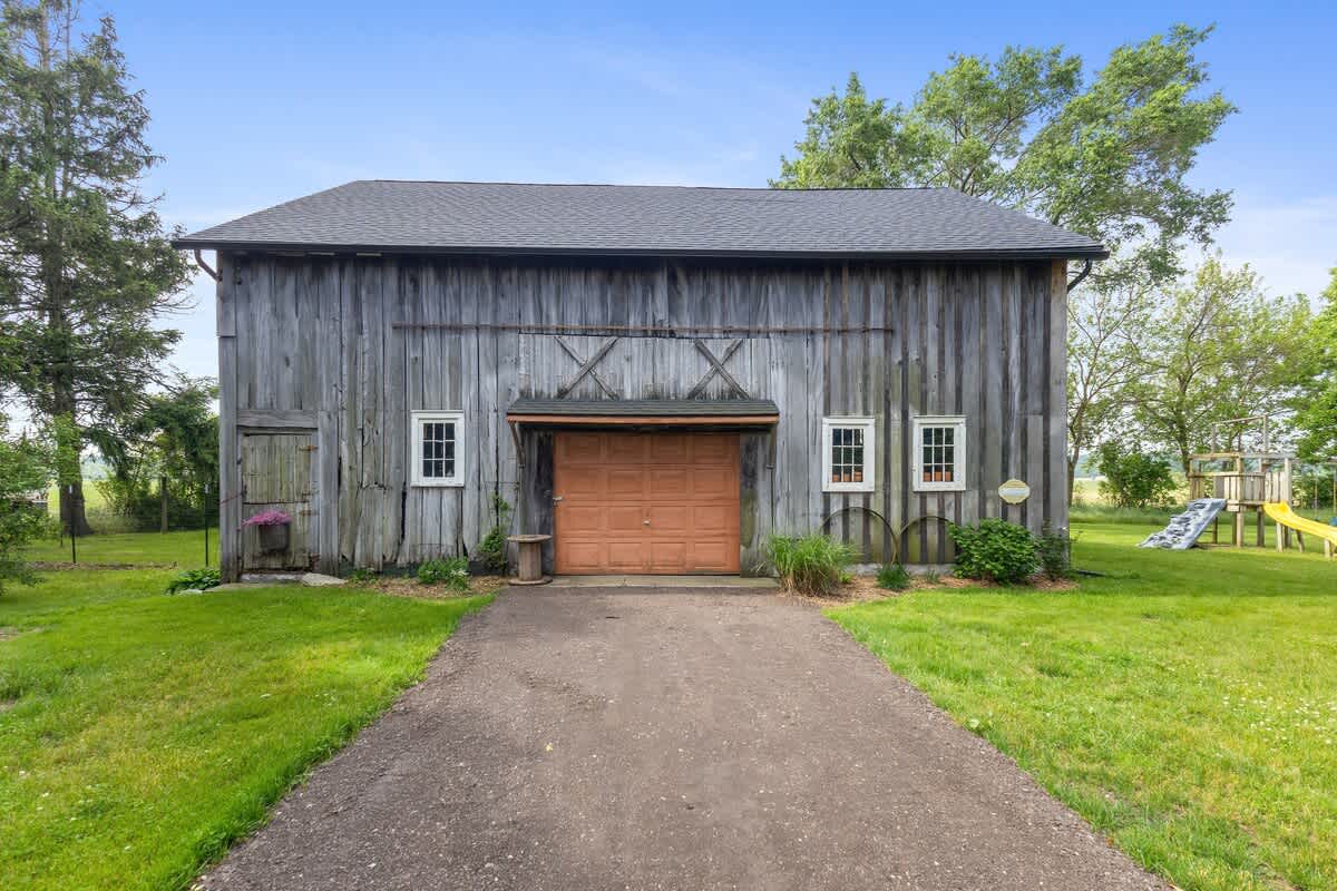 Rustic wooden barn with a large orange garage door and several windows, set against a backdrop of green grass and trees. A gravel driveway leads up to the barn.
