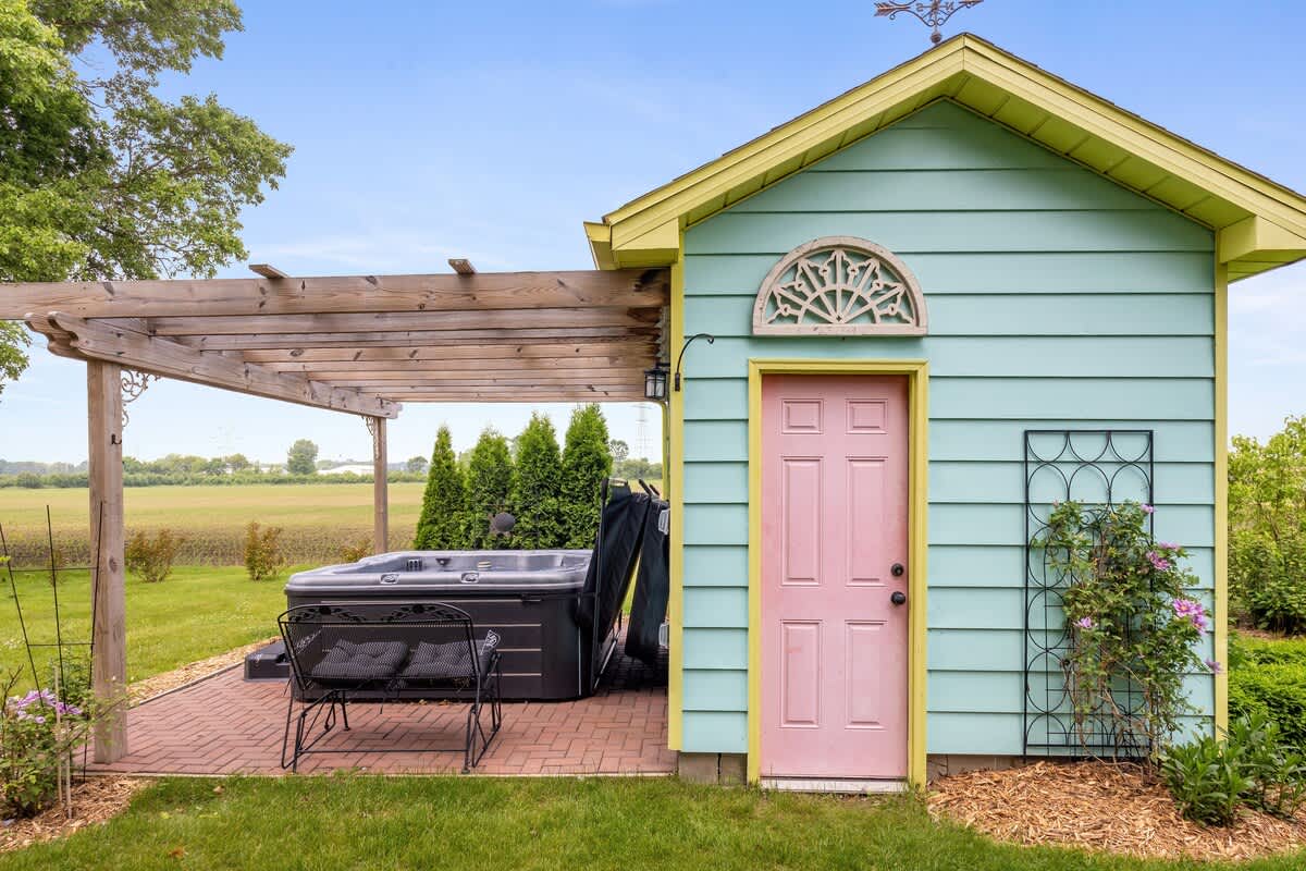 A covered hot tub sits on a brick patio under a wooden pergola, adjacent to a charming light blue shed with a yellow trim and a pink door.
