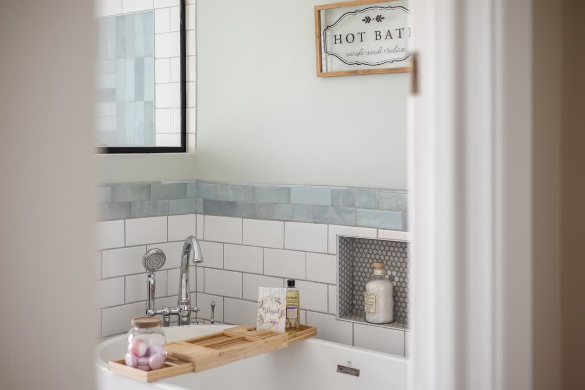Close-up of a white freestanding bathtub with a wooden caddy holding bath items, set against white subway tiles and light blue accent tiles. A framed "HOT BATH" sign hangs above.