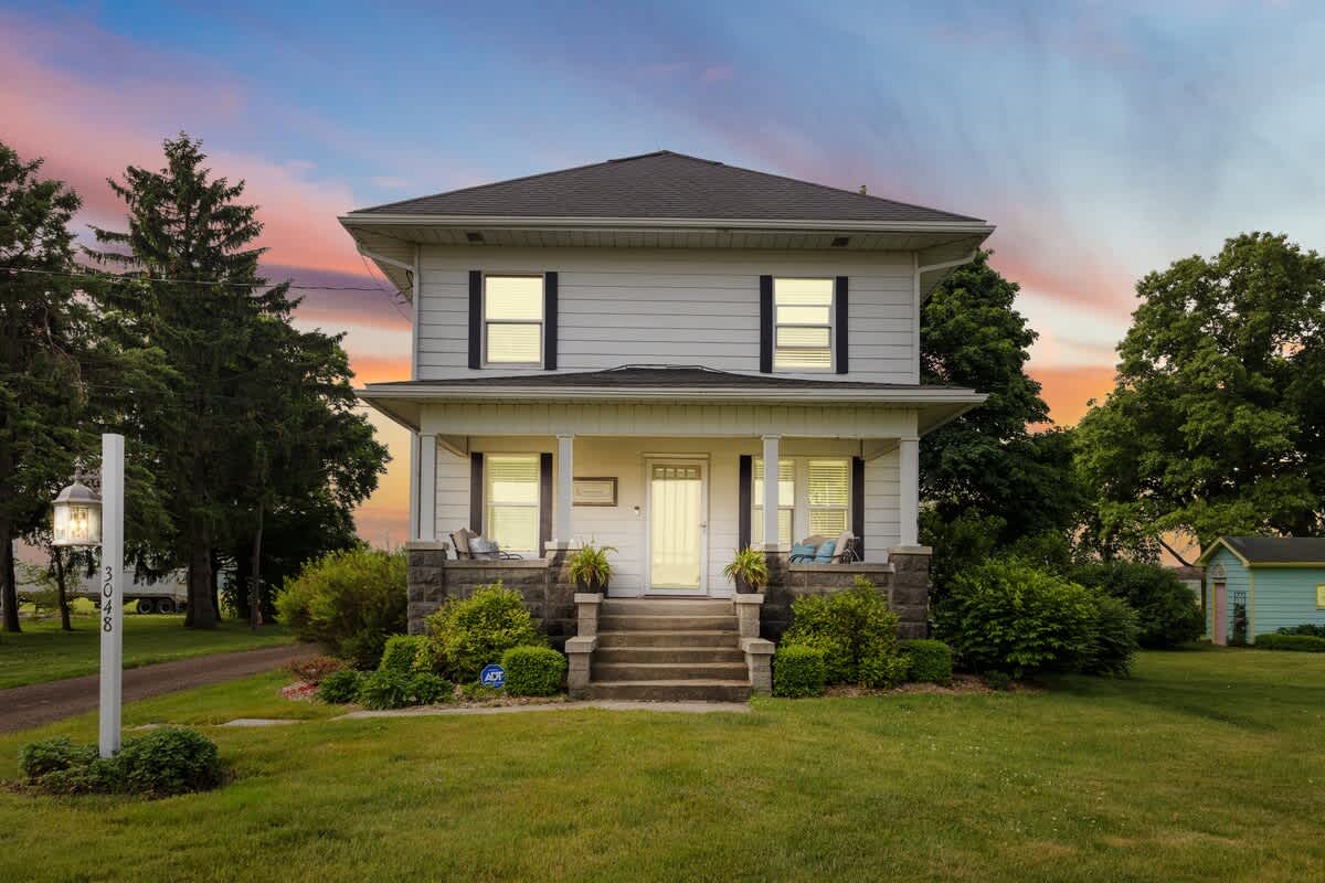 The classic white farmhouse features a welcoming front porch with stone pillars and concrete steps leading to the main entrance. Lush green bushes and a well-kept lawn frame the property under a colorful sunset sky.