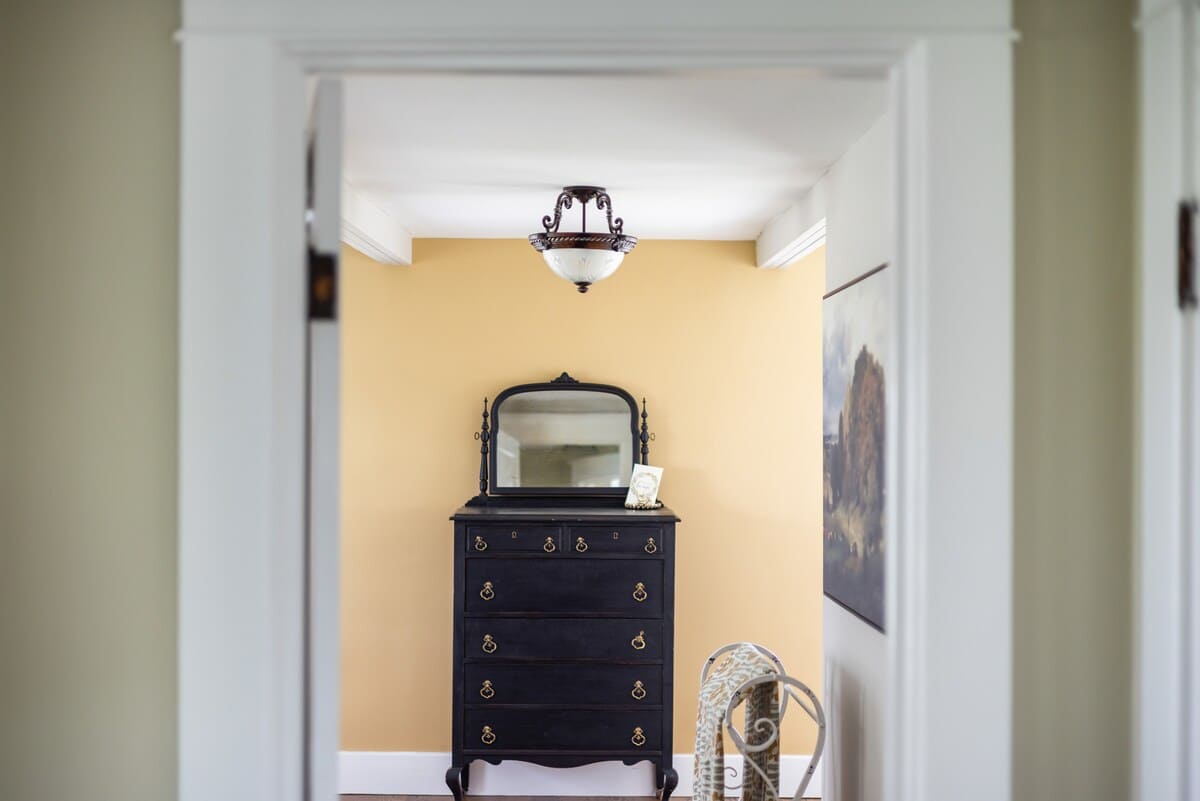 A narrow hallway with light yellow walls features a dark wooden dresser with a matching mirror, illuminated by an ornate ceiling light fixture, leading into another room.
