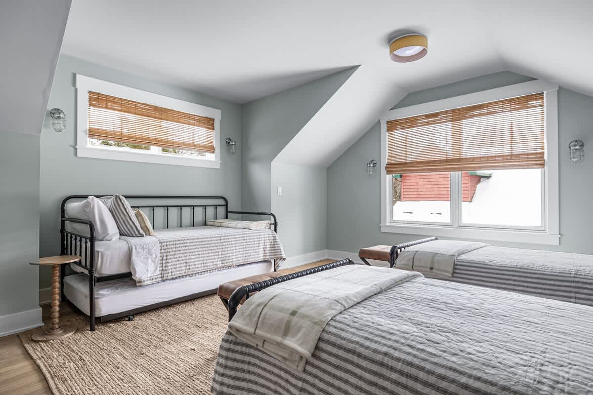 Bright loft bedroom with a daybed and two twin beds, all featuring striped bedding. The room has light blue walls, sloped ceilings, and windows with bamboo blinds, complemented by a natural fiber rug.