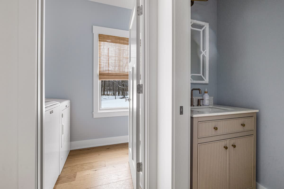 Laundry area with a white washer and dryer, adjacent to a small bathroom vanity with a ribbed cabinet and white countertop. The room features light blue walls and hardwood floors.