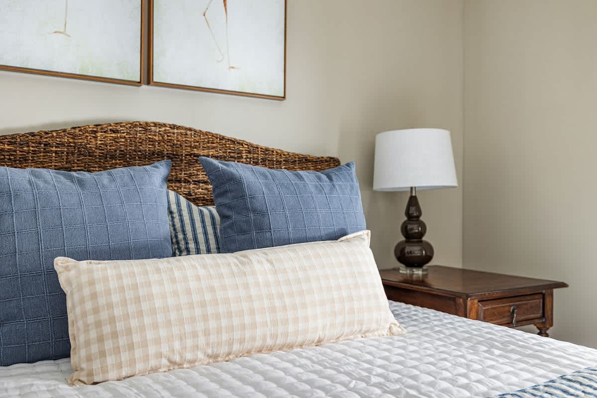 Cozy bedroom featuring a woven wicker headboard, blue and plaid throw pillows, and a white quilted comforter. A dark wood nightstand with a white lamp sits beside the bed.