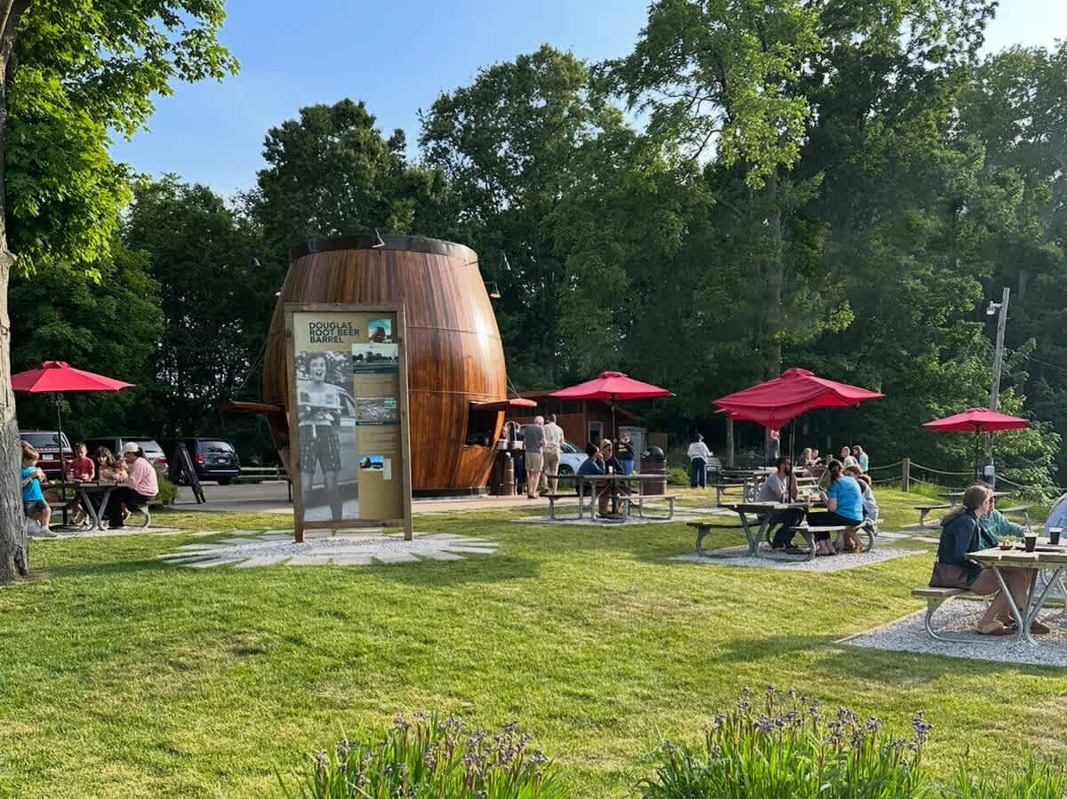An outdoor dining area features several picnic tables with red umbrellas, set on a grassy lawn next to a unique barrel-shaped building. People are enjoying food and drinks in the sunny weather.
