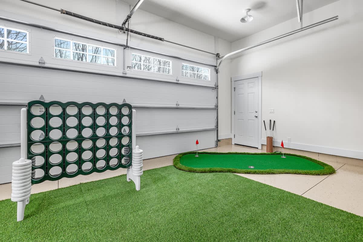 A corner of the game room, located in a garage, featuring a green artificial turf putting green with a red flag, and a large white and green Connect Four game set against a white garage door.
