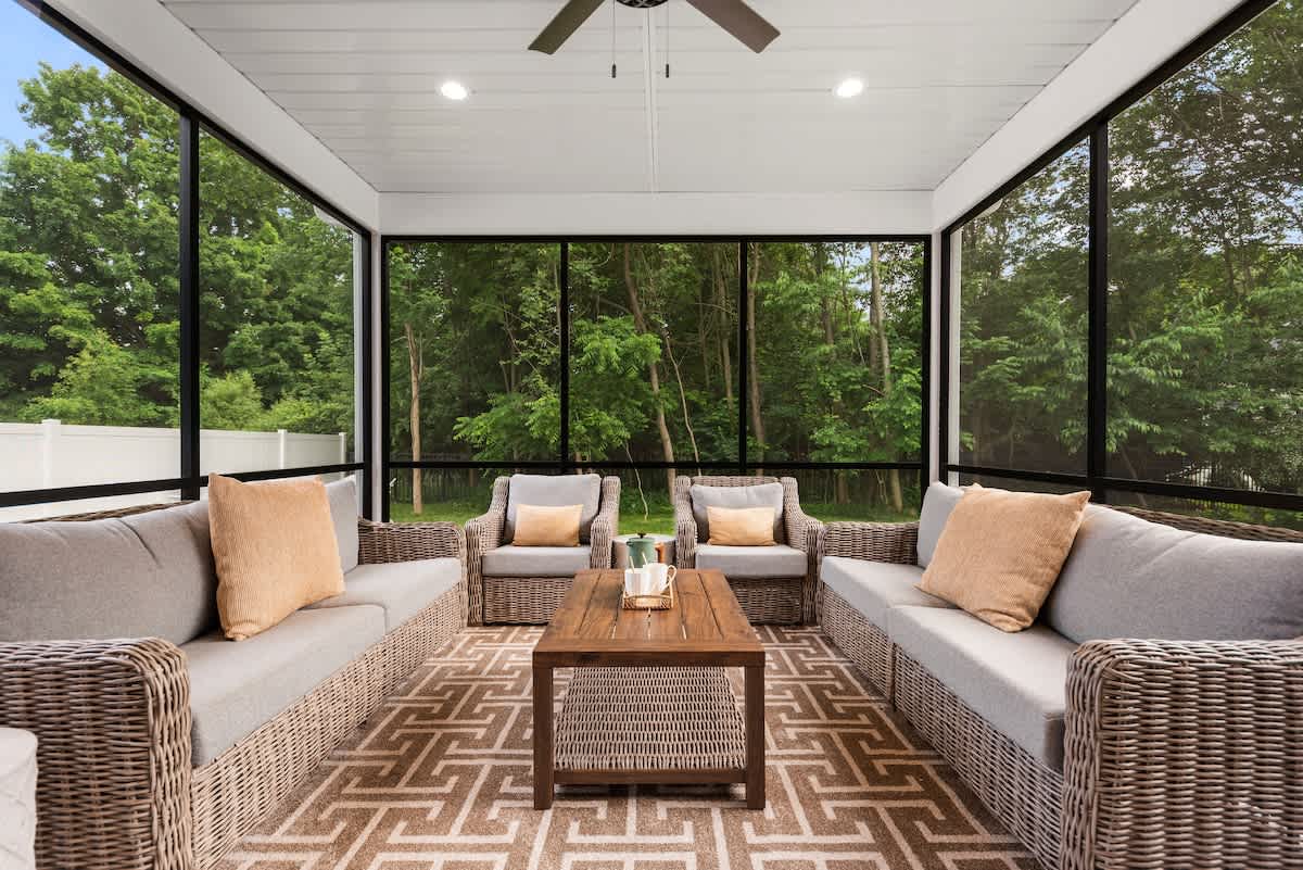 Spacious screened porch featuring two wicker sofas and two armchairs with light gray cushions and tan throw pillows, arranged around a rustic wooden coffee table on a geometric patterned rug. The porch offers expansive views of a lush green forest through large black-framed screens.