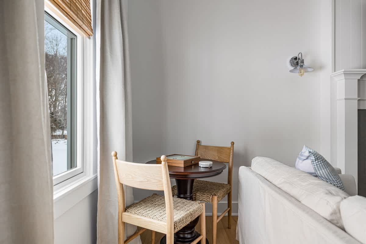 A small, cozy nook in the living room features a round wooden table with two light-colored wooden chairs with woven seats. A window with light drapes and a bamboo blind offers a glimpse of a snowy outdoor landscape.